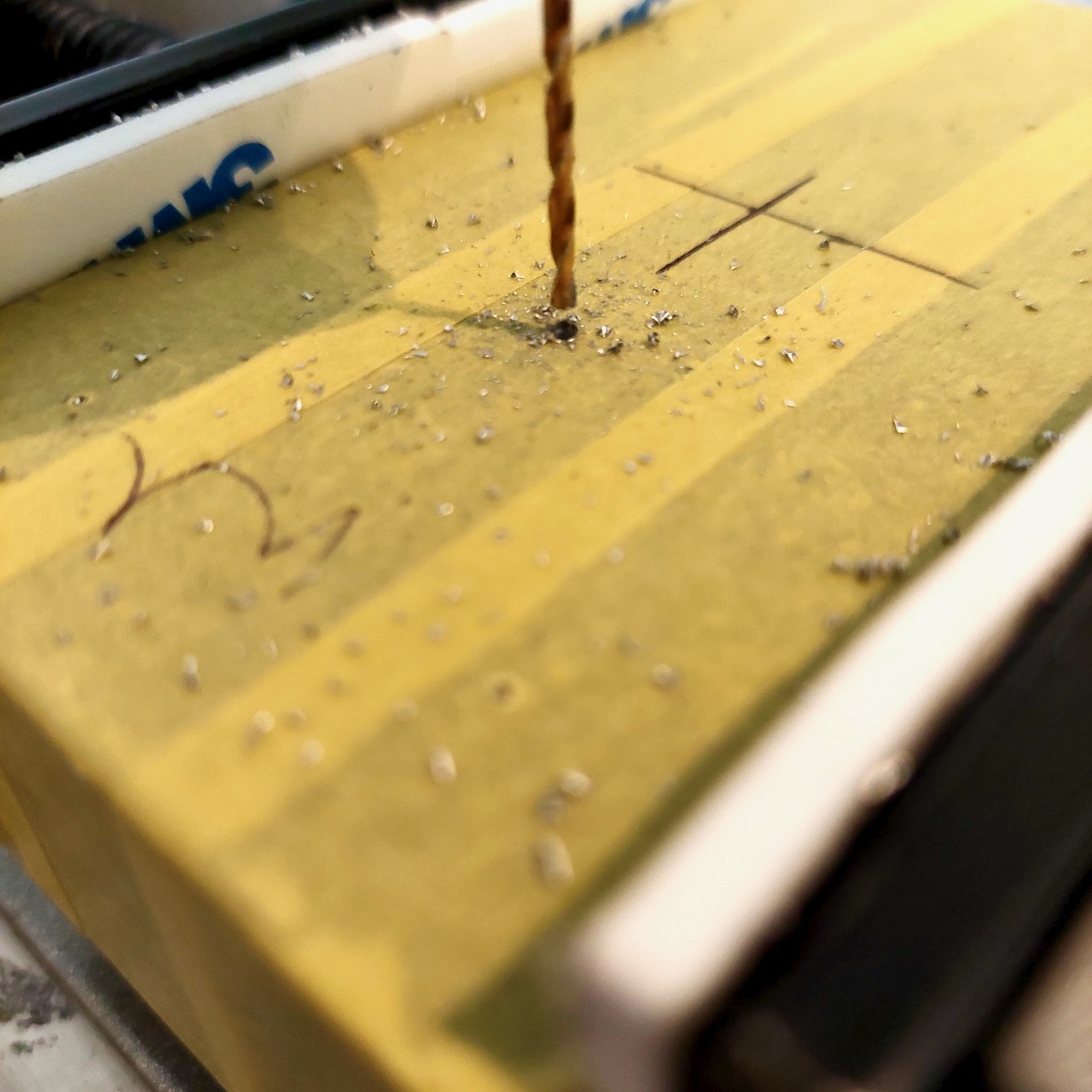A fine twist bit cutting a pilot hole into a masked enclosure, with aluminium swarf across the tape.