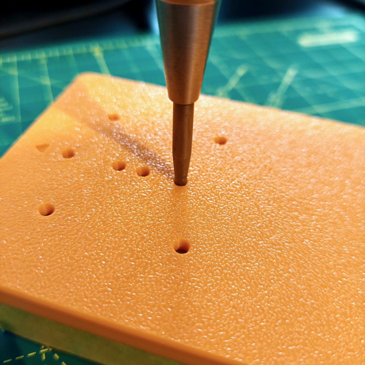 A centre punch resting in one of the bores of the drill jig, with more bores across the plate around it.