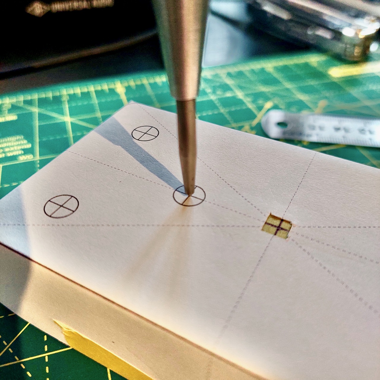 A centre punch resting on one of the printed crosshairs, on a template taped over an enclosure.