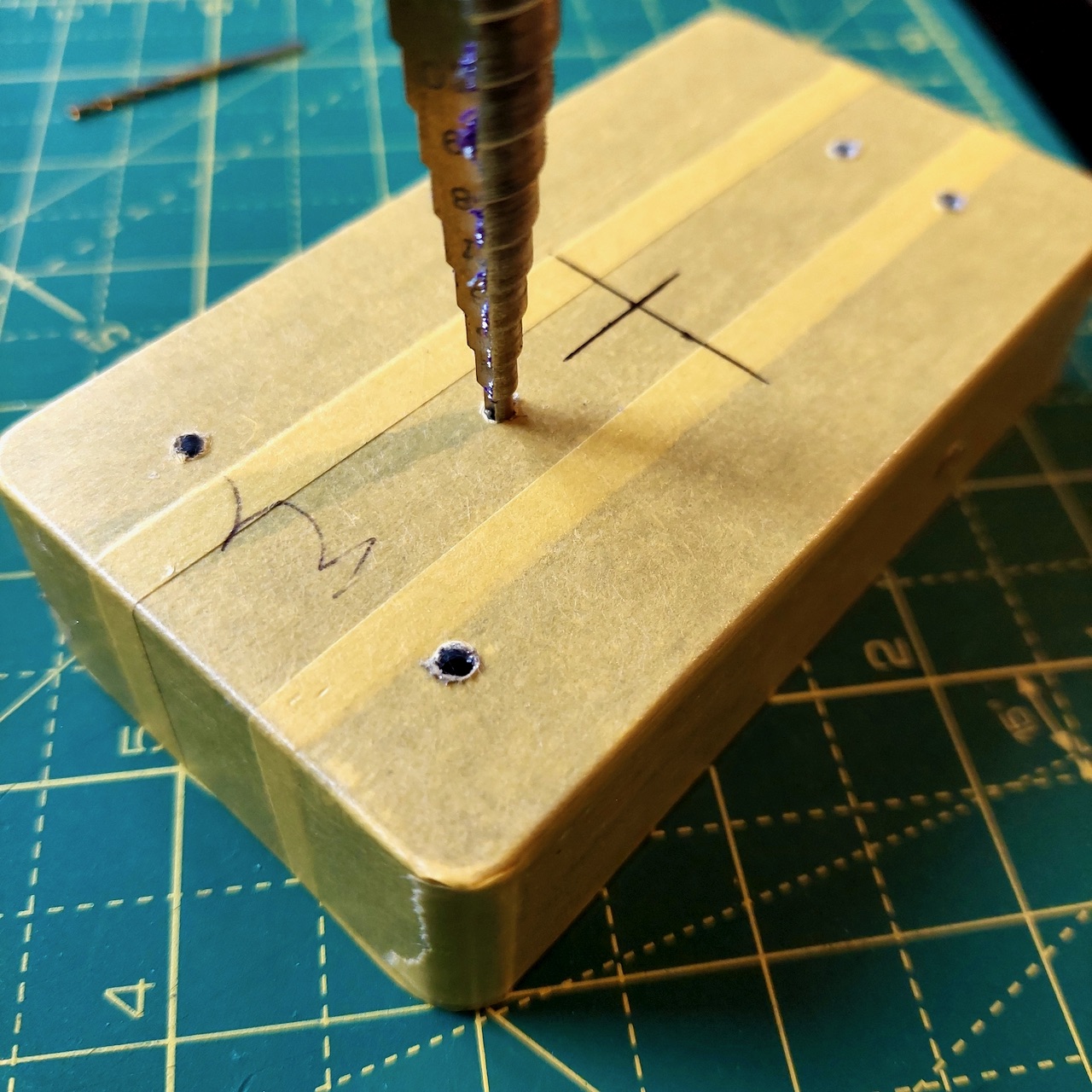 A step drill bit poised over a pilot hole in a masked enclosure, with more drilled holes around it.